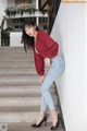 A woman in a red blouse and jeans leaning against a wall.