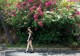 A woman walking down the street in front of a bush of flowers.